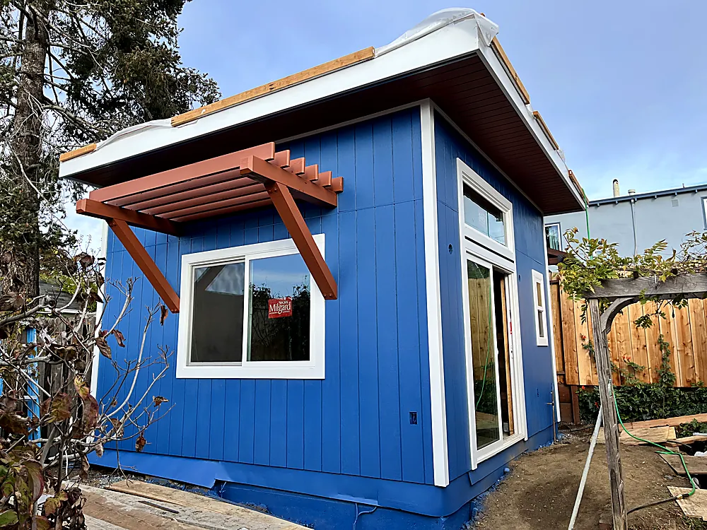 A detached backyard studio painted blue with white trim and a cedar bracket